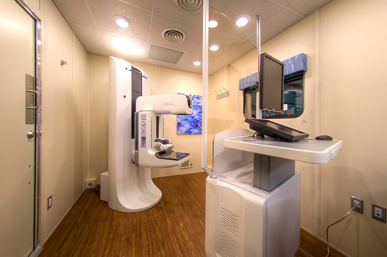 Medical imaging room equipped with a state-of-the-art mammography machine on the left and a computer workstation to the right, featuring wood flooring and neutral-colored walls.
