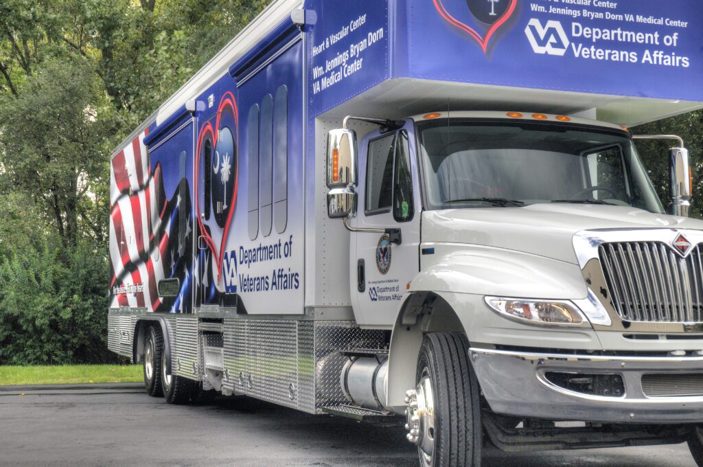 A large Department of Veterans Affairs mobile medical unit is parked on pavement, featuring patriotic graphics and VA branding on its side and front. This specialized unit offers mammography services to veterans, bringing essential women’s health screenings directly to the community.