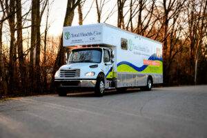 A white Total Health Care mobile clinic truck is parked on a paved road, providing essential healthcare services with leafless trees in the background.