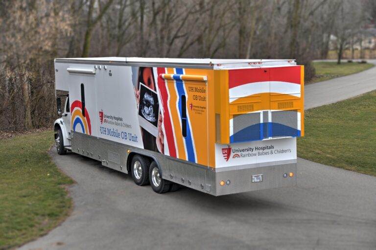 A large University Hospitals Rainbow Babies & Children’s mobile OB unit truck driving on a paved road surrounded by trees.