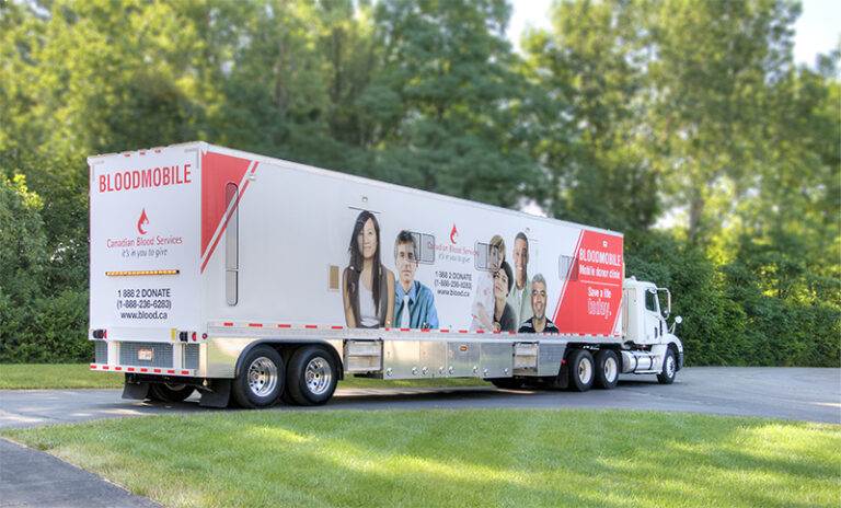 A white Canadian Blood Services bloodmobile truck is parked on a paved area beside grass and trees, displaying promotional images and donation information on its side.