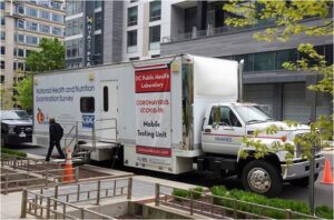 A mobile health COVID-19 testing unit truck is parked on a city street near buildings, with a person standing next to it.