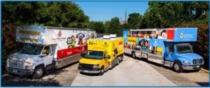 Three colorful mobile clinics are parked side by side outdoors on a driveway, surrounded by trees.