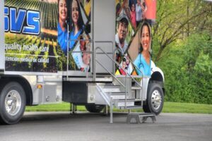Mobile health clinic truck with metal stairs and ramp access, parked on pavement near green trees. The truck displays photos of diverse people and healthcare workers.