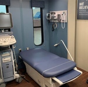 A medical examination room with an exam table, ultrasound machine, wall-mounted diagnostic tools, and a window.