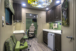 Interior of a modern mobile medical clinic with green accents, white countertops, cabinets, chairs, and a sky-themed ceiling light, designed to support continuity of care in a comfortable setting.
