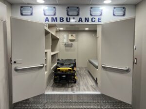 Interior view of an ambulance with open double doors, showing a stretcher, shelves on the left, and a bench on the right, designed to support continuity of care during patient transport.