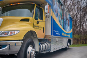 A large yellow and blue medical simulation training truck, supporting continuity of care, is parked on a paved road next to trees.