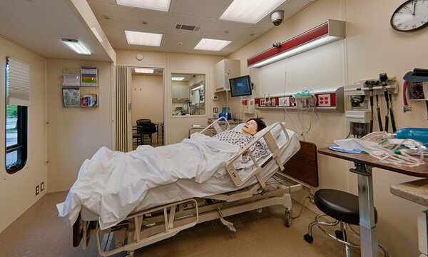 A medical simulation mannequin lies in a hospital bed inside a clinical training room equipped with medical devices and monitors, providing realistic training for continuity of care.