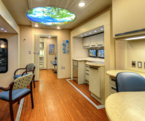 Modern medical facility room with wood flooring, beige walls, chairs, cabinets, and a ceiling light fixture displaying an image of tree branches and sky—a testament to CMS rural health investment in patient-centered spaces.
