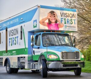 A large Vision First mobile health clinic eye truck with Cleveland Clinic branding is parked outdoors; a child making glasses with her hands is pictured on the truck's sign.
