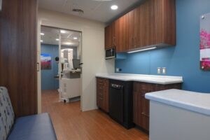 Small medical office kitchenette with wood cabinets, blue walls, a dishwasher, and a bench. Medical imaging equipment is visible in an adjacent room, supporting mobile behavioral health services offered on-site.