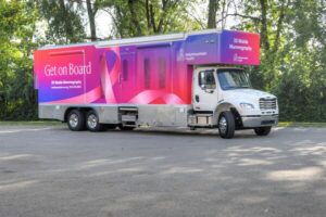 A large mobile mammography truck with pink and purple branding from Intermountain Health, also supporting mobile behavioral health services, is parked on pavement near trees.