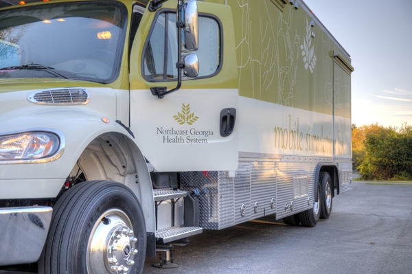 A Northeast Georgia Health System mobile medical unit is parked on a paved surface, with trees and sky visible in the background.
