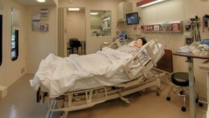 A medical mannequin lies in a hospital bed in a simulation room equipped with medical devices, monitors, and various supplies.