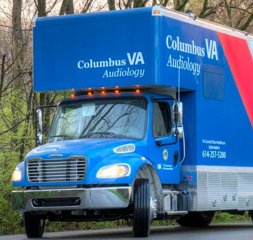 Blue Columbus VA Audiology mobile clinic truck parked on a road, surrounded by trees. The truck, reminiscent of advanced mobile simulation laboratories, displays contact information and VA branding.