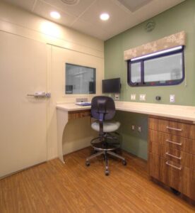 A small office with a desk, swivel chair, computer monitor, window, and cabinets. The room has wood flooring and green and beige walls, creating an organized space ideal for coordinating mobile simulation laboratories.