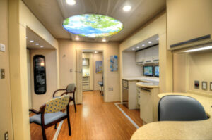 Interior of a mobile audiology clinic featuring seating, countertops, medical equipment, and a ceiling light panel depicting a sky view.